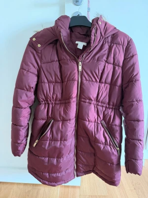 Mammajacka- Vinröd pufferjacka med huva M - Säljer en vinröd mamma pufferjacka med huva och dragkedja framtill. Jackan har två snedställda fickor med dragkedja och guldiga detaljer. Quiltad design och lätt insvängd midja för snygg passform. Perfekt för kalla vinterdagar.