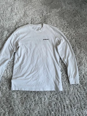 Vit långärmad Patagonia t-shirt - Vit långärmad t-shirt från Patagonia med liten logga på bröstet och stor färgglad logga med bergmotiv på ryggen. Tillverkad i mjuk bomull och har en klassisk passform med rund hals.