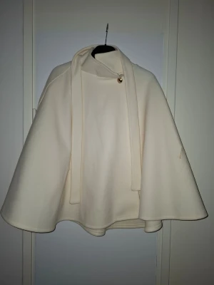 Krämvit cape med knytband - Stilren cape i krämvitt tyg med hög krage och långa knytband framtill. Kappan har en lös, svepande passform och stängs med en dekorativ knapp vid halsen. Perfekt för dig som vill ha en elegant och unik look. Finns nån liten fläck men går bort i tvätten.