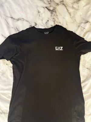 Svart EA7 Emporio Armani t-shirt - Snygg svart t-shirt från EA7 Emporio Armani med vit logga tryckt på bröstet. Klassisk rund halsringning och korta ärmar. Tillverkad i mjukt material som känns skönt mot huden. Perfekt för en clean och stilren look.