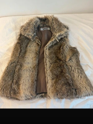 Brun fluffig fuskpälsväst - Säljer en snygg och fluffig brun fuskpälsväst med bred krage och mjukt foder. Västen har en naturlig färgskiftning i brunt och beige som ger en cool look. Perfekt att slänga över en hoodie eller tröja för extra stilpoäng.