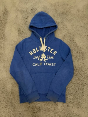 Blå Hollister hoodie med tryck - Blå hoodie från Hollister med vit text 'Surf Fest Calif Coast' och ett lägereldstryck på bröstet. Hoodien har vita dragsnören, stor känguruficka framtill och ribbade muddar vid ärmar och nederkant. Perfekt för chill dagar och streetwear. ⚠️⚠️tröjan är storlek L men har krympt till M, passar även liten L, den är också smutsig vid arm sluten men går att tvätta⚠️⚠️