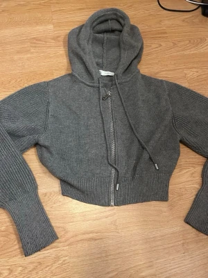 Grå cropped stickad hoodie - Säljer en grå cropped hoodie i stickat material med dragkedja framtill och ribbade muddar på ärmar och nederkant. Hoodien har en stor huva och dragsko. Perfekt för en chill och avslappnad stil.