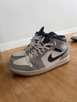 Nike Air Jordan 1 Mid grå/vit - Snygga Nike Air Jordan 1 Mid sneakers i grått, vitt och svart. Klassisk design med svart swoosh, vita paneler och gråa overlays. Skorna har högt skaft och är tillverkade i läder med textilfoder. Perfekta för dig som gillar streetwear och ikoniska sneakers.