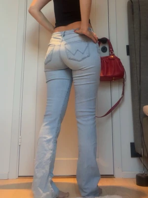 Wrangler bootcut jeans - Säljer ett par ljusblå bootcut jeans med klassiska fickor och snygga sömdetaljer bak. Jeansen har hög midja och är tillverkade i ett stretchigt denimtyg som sitter tajt över höfterna och låren men släpper ut nedtill.
