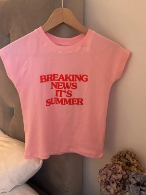 Rosa t-shirt - Säljer en ljusrosa t-shirt med trycket 'BREAKING NEWS IT'S SUMMER' i rött framtill. T-shirten har korta ärmar och en klassisk rund halsringning.  Väldigt skön i material 