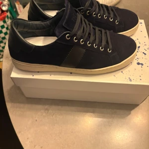 Mörkblå sneakers från The Gallerist - Snygga mörkblå sneakers från The Gallerist med ovandel i mocka och detaljer i läder. Skorna har vita sulor, svarta skosnören och metallfärgade öljetter. Perfekta för en stilren och modern look. Storlek 44. De är använda 2 gånger så i väldigt bra skick som nya.