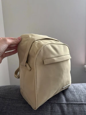 Beige miniryggsäck i nylon - Snygg beige miniryggsäck med minimalistisk design. Tillverkad i slätt nylonmaterial med justerbara axelband och dragkedja baktill. Perfekt för dig som vill ha en stilren och enkel accessoar till vardagslooken. Mått 26x20x10 cm 