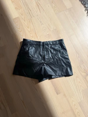 Svarta shorts i skinnimitation - Snygga svarta shorts i skinnimitation med normal passform och midja. Klassisk design med fickor fram och hällor för bälte. Perfekta för att lyfta vilken outfit som helst.