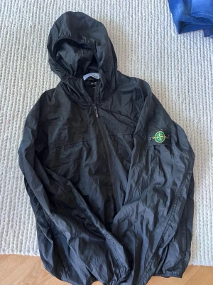 Svart vindjacka från Stone Island - Säljer en svart vindjacka från Stone Island med klassisk patch på ärmen. Jackan har huva, dragkedja framtill och är tillverkad i lätt, vindtätt material. Perfekt för dig som gillar streetwear och vill ha en stilren look.