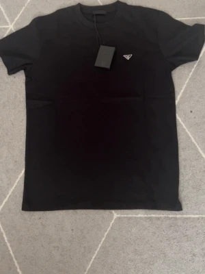Svart t-shirt från Prada - Snygg svart t-shirt från Prada med klassisk passform och ikonisk metallogo på bröstet. Tillverkad i mjuk bomull som känns skön mot huden. Perfekt för dig som gillar stilrena och exklusiva plagg.