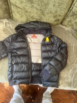 Parajumpers SHEEN pufferjacka - Snygg svart pufferjacka från Peak Performance med huva och dragkedja. Jackan har quiltad design, ribbade muddar och praktiska innerfickor. Insidan är ljusgrå och materialet är glansigt och lätt, perfekt för kalla vinterdagar. Kom med bud vi löser något😉
