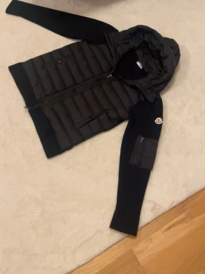 Svart dunväst med huva och ärmar - Snygg svart dunväst med vadderad framsida, stickade svarta ärmar och huva. Dragkedja framtill och fickor med dragkedja. Moncler-logga på ena ärmen och extra ficka på ärmen. Perfekt för lager-på-lager-stil.