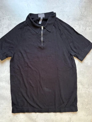 Svart polotopp med dragkedja H&M - Svart polotopp från H&M i muscle fit-modell. Toppen har korta ärmar, klassisk krage och en snygg dragkedja framtill. Perfekt för en clean och stilren look. Materialet känns mjukt och bekvämt mot huden.