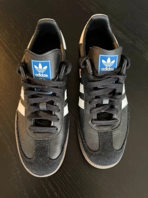 Adidas Samba svartvita sneakers - helt oanvända (felköp) - Klassiska Adidas Samba sneakers i svart med vita ränder och blå logga på plösen. Skorna har lågt skaft, svart mocka och läderdetaljer samt en brun gummisula. Perfekta för dig som gillar retrostil och streetwear.