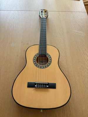 Klassisk akustisk gitarr i trä - Säljer en klassisk akustisk gitarr som är i barn storlek med naturfärgad framsida och mörkbrun baksida.  Perfekt för dig som vill börja spela eller bara jamma hemma nästa hel oanvänd och inga tecken på skador och vätskan ingå 