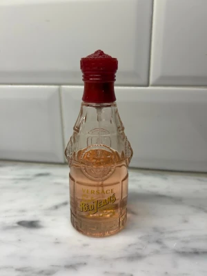 Versace Red Jeans parfymflaska - Versace Red Jeans parfym i en unik, genomskinlig glasflaska med detaljerad relief och röd kork. Flaskan har en ljusrosa tonad vätska och guldfärgad text på framsidan. Perfekt för dig som gillar klassiska och ikoniska dofter.