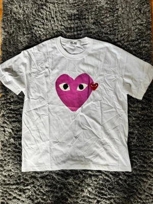 Vit t-shirt CDG - Vit CDG t-shirt med ett stort rosa hjärta med ögon och ett litet rött hjärta på bröstet. Klassisk rund hals och korta ärmar. Perfekt för dig som gillar lekfulla och unika prints.