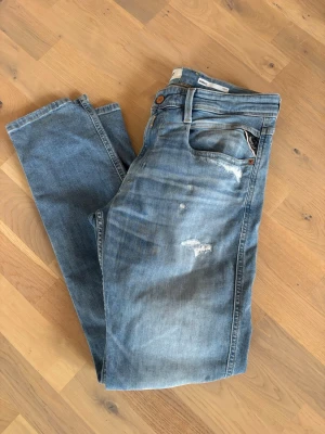 Blå slitna Replay jeans med raka ben - Säljer ett par ljusblå jeans med slitningar och små hål på låret och knät. Jeansen har klassisk femficksmodell, normal midja och raka ben. Materialet är denim i bomull och de har en avslappnad, trendig look.