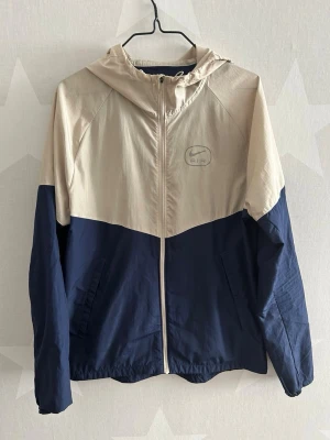 Nike Air vindjacka blå/beige - Snygg tvåfärgad vindjacka från Nike Air i beige och marinblått. Jackan har huva, dragkedja framtill, två sidofickor och stor Nike Air-logga i rött på ryggen. Tillverkad i lätt och vindtätt material, perfekt för blåsiga dagar.