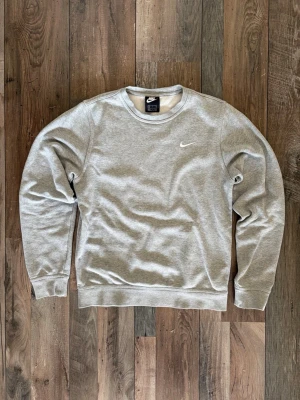 Grå sweatshirt från Nike - Klassisk grå sweatshirt från Nike med rund halsringning och vit broderad Swoosh-logga på bröstet. Tröjan har ribbade muddar vid ärmslut och nederkant. Perfekt för en avslappnad och sportig stil.
