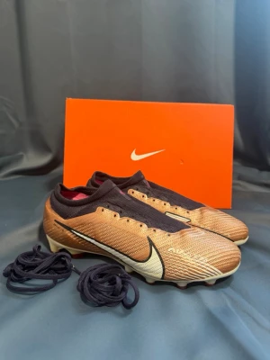 Nike Mercurial vapor 15 elite  - Snygga Nike Mercurial fotbollsskor i brons med svart och vitt. Skorna har en strömlinjeformad design, stora AIR-detaljer på sidan och utbytbara svarta skosnören. Ovandelen är i syntetmaterial och sulan har dobbar för bra grepp på planen.