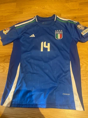 Italien matchtröja Chiesa #14 blå - Säljer en blå Italien matchtröja med Chiesa och nummer 14 på ryggen. Tröjan har korta ärmar, vita detaljer på sidorna och officiella märken på bröstet och ärmarna. Tillverkad i funktionsmaterial med AEROREADY-teknologi.