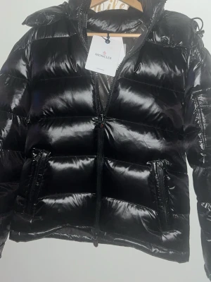 Svart dunjacka från Moncler - Snygg svart dunjacka från Moncler med glansig finish, huva och dragkedja framtill. Jackan har två stora fickor med dragkedja och Moncler-logga på armen. Perfekt för kalla vinterdagar och riktigt trendig look
