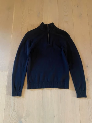 Mörkblå stickad tröja med halvzip - Stilren mörkblå tröja med hög krage och halv dragkedja framtill. Tröjan har långa ärmar och ribbade muddar vid ärmslut och nederkant. Perfekt för lager på lager och enkel att matcha med olika stilar.