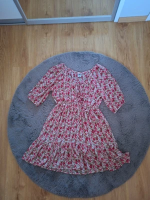 Blommig kortklänning från Isolde - Supersöt blommig klänning från Isolde i storlek 42. Klänningen har en vit bas med röda och gröna blommor, trekvartsärm och volang nertill. Perfekt för vår och sommar med sin lätta och luftiga känsla.