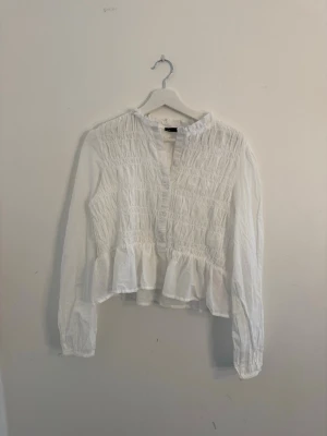 Vit smockad blus med volang - Söt vit blus med smockad överdel och volang nedtill. Blusen har lång ärm och liten ståkrage, samt en avslappnad passform. Perfekt att styla med jeans eller kjol för en trendig look.