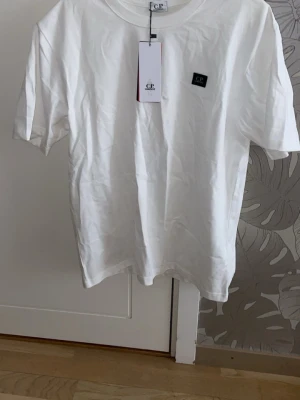 Vit t-shirt från C.P. Company - Vit t-shirt från C.P. Company med klassisk passform och korta ärmar. T-shirten har en liten svart logotyp på bröstet och är tillverkad i mjuk bomull. Perfekt för en clean och stilren look.