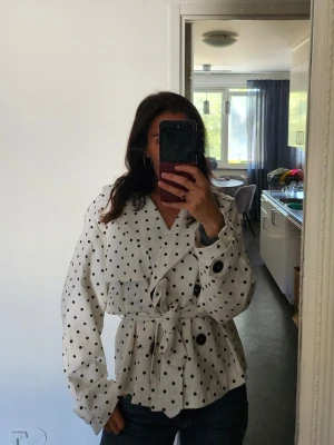 Vit trenchcoat med svarta prickar - Snygg vit trenchcoat med svarta prickar och knytskärp i midjan. Jackan har dubbelknäppning framtill, stora svarta knappar och klassisk krage. Materialet ser ut att vara bomull eller en bomullsblandning. Perfekt för dig som vill sticka ut med en trendig och lekfull look.