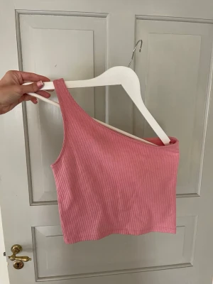 Rosa oneshoulder topp från Gina Tricot - Säljer en ribbad rosa oneshoulder topp från Gina Tricot. Toppen är croppad och har en snygg asymmetrisk axelbandslös design som ger en trendig look. Perfekt att matcha med höga jeans eller kjol för en cool stil.