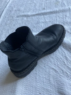 Svarta stövletter från Rieker - Säljer ett par svarta stövletter från Rieker i skinnimitation med grov sula och dragkedja på sidan. Skorna har en chunky look och kontrastsömmar runt sulan. Perfekta för dig som vill ha en cool och bekväm sko till hösten eller vintern.