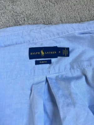 Ljusblå skjorta från Ralph Lauren - Klassisk ljusblå skjorta från Ralph Lauren i slim fit-modell. Skjortan har långa ärmar, knappar framtill och den ikoniska broderade loggan på bröstet. Tillverkad i mjuk bomull och passar perfekt till en stilren look.