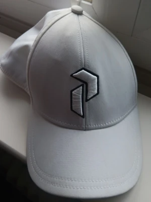 Vit keps med svart broderad logga - Snygg vit keps med böjd skärm och svart broderad logotyp framtill samt mindre logga på sidan. Kepsen har klassisk passform och är tillverkad i ett mjukt tygmaterial. Perfekt accessoar för en clean och sportig stil.