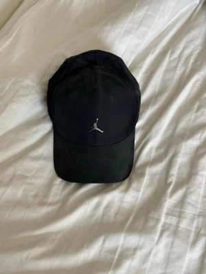 Svart Jordan keps med broderad logga - Snygg svart keps från Jordan med den klassiska Jumpman-loggan broderad framtill i silver. Justerbar rem baktill för perfekt passform. Tillverkad i ett slitstarkt tyg som passar till alla streetwear-outfits.