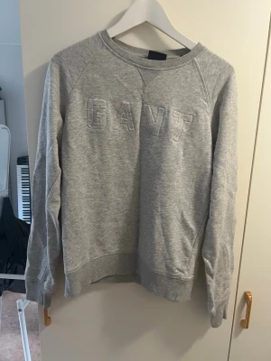 Grå sweatshirt från GANT - Säljer en klassisk grå sweatshirt från GANT med broderad logga framtill. Tröjan har rund halsringning, långa ärmar och ribbade muddar vid ärmslut och nederkant. Perfekt för en chill och avslappnad stil.