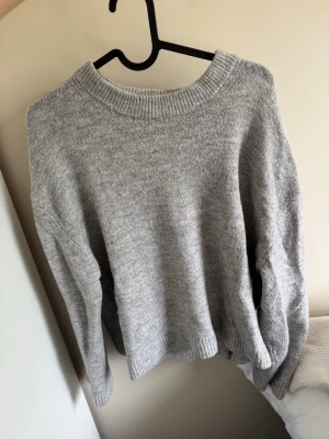Grå stickad tröja med rund hals - Mysig grå stickad tröja med rund halsringning och långa ärmar. Tröjan har ribbstickade muddar vid ärmslut och nederkant, vilket ger en klassisk och enkel look. Perfekt för lager på lager under kyliga dagar. Lite urtvättad och nopprig. 