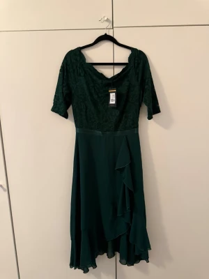 Mörkgrön långklänning med spets - Elegant mörkgrön långklänning med spetsdetaljer upptill och volang längs kjolen. Klänningen har v-ringning, halvlånga ärmar och ett flytande, böljande tyg i kjolen. Perfekt för speciella tillfällen när du vill känna dig extra fin.