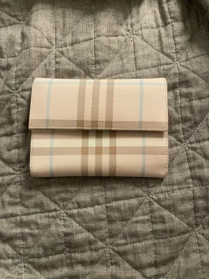 plånbok från Burberry - Snygg plånbok från Burberry i klassiskt beige rutmönster med ljusblå detaljer. Insidan är ljusrosa med flera kortfack, myntfack med dragkedja och knappstängning. Perfekt för dig som gillar stilrena accessoarer med lyxig känsla. Fick den av en kollega. 