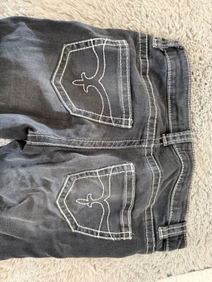 Grå jeans med vita sömmar - Säljer ett par grå jeans med tydliga vita kontrastsömmar och unika broderade bakfickor. Klassisk femficksmodell med dragkedja och knappstängning. Jeansen har en avslappnad passform och är tillverkade i denim. Från Gina i storlek 158. 