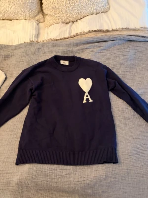 Mörkblå stickad tröja med hjärtdetalj - Mörkblå stickad tröja med rund halsringning och ribbade muddar. På bröstet finns en vit hjärtdetalj med bokstaven A under. Tröjan har en klassisk passform och är perfekt för dig som gillar stilrena detaljer.