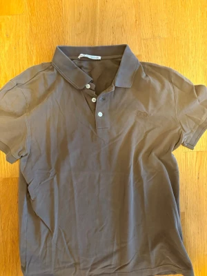 Brun pikétröja från Tiger of Sweden - Snygg brun pikétröja från Tiger of Sweden med klassisk krage och knappar framtill. Tröjan har korta ärmar och diskret broderad logga på bröstet. Perfekt till jeans eller chinos för en clean look.
