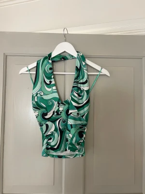 Grön mönstrad halterneck topp - Säljer en grön och vit halterneck topp med djup v-ringning och swirl-mönster. Toppen har smala axelband och är croppad, perfekt för en trendig look. Materialet känns mjukt och stretchigt, troligen polyester eller syntet.