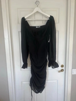 Svart omlottklänning med volang - Snygg svart klänning med långa transparenta ärmar, omlottdesign och volangdetaljer. Klänningen har snörning framtill och rynkade detaljer längs kjolen för en trendig look. Perfekt för dig som vill sticka ut.