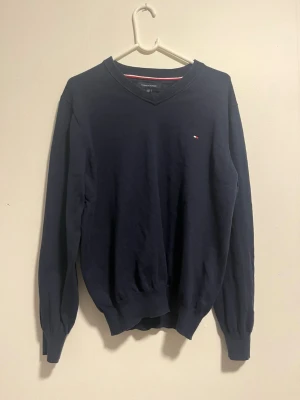 Marinblå tröja från Tommy Hilfiger - Klassisk marinblå tröja från Tommy Hilfiger med v-ringad hals och diskret logga på bröstet. Tröjan har långa ärmar och ribbade muddar vid ärmslut och nederkant. Perfekt till en stilren vardagslook.