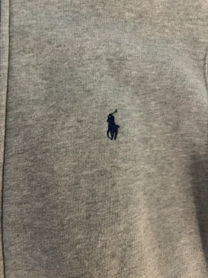 Grå zip hoodie från Polo Ralph Lauren - Snygg grå hoodie med dragkedja från Polo Ralph Lauren. Klassisk design med mörkblå logga broderad på bröstet, huva och två stora fickor framtill. Tillverkad i mjukt bomullsmaterial som är skönt att bära. Aldrig använd. Passar 14-16 år. 
