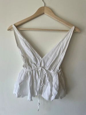 Vit linnetopp från Brandy Melville - Supersöt vit linnetopp från Brandy Melville med djup v-ringning och knyt i midjan. Toppen är ärmlös och har en luftig, flowy passform – perfekt för varma dagar. Tillverkad i mjukt bomullstyg.